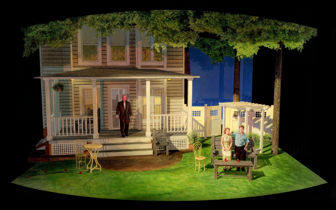 A man on a porch and a couple on a bench are in a theater set resembling a house with trees. The scene is lit warmly, evoking calm.