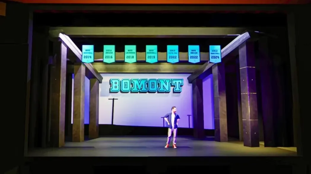 Man on stage under a 'BOMONT' sign and sports banners. Blue and purple lighting sets a dramatic mood with stone-like pillars flanking the scene.
