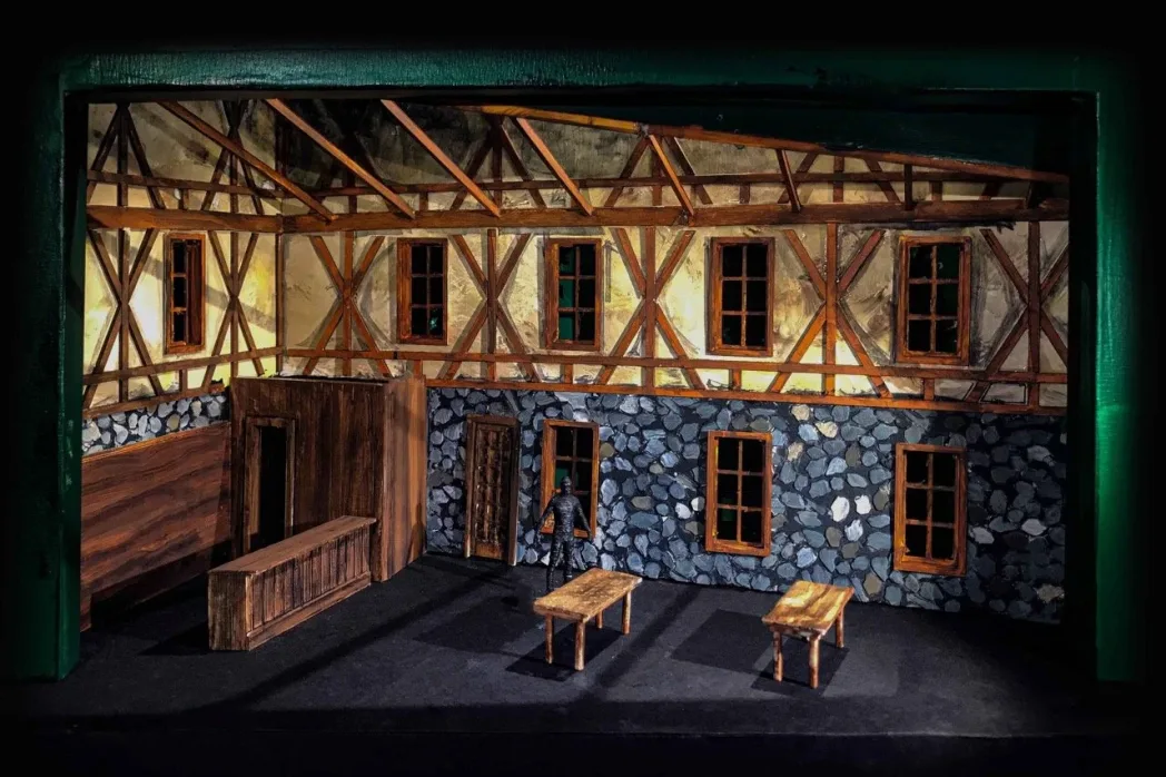 Stage set with rustic timber-framed walls and stone accents. A lone person stands near a window, with wooden tables on a wooden floor.