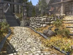 Stone path leads to a medieval village with wooden houses; flowers and greenery surround the scene on a peaceful, sunny day.