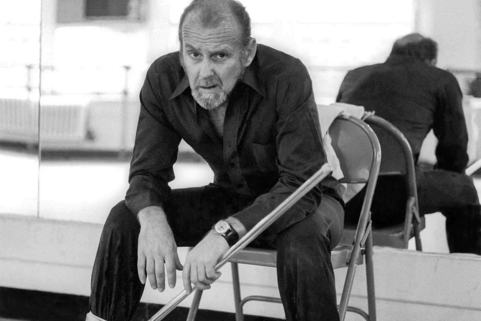 Bob Fosse seated in a mirrored studio with a cane.